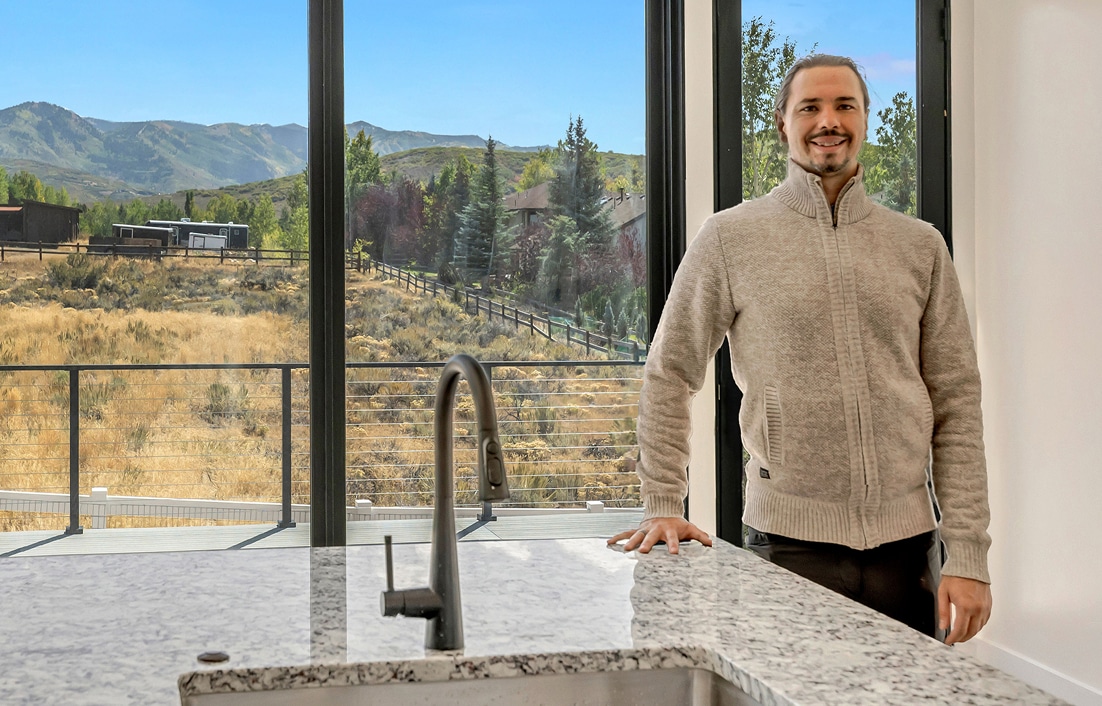 Man standing in a modern kitchen with granite countertop, smiling, showcasing large windows with a mountain view, highlighting Oak-n-Crete's custom home building focus.