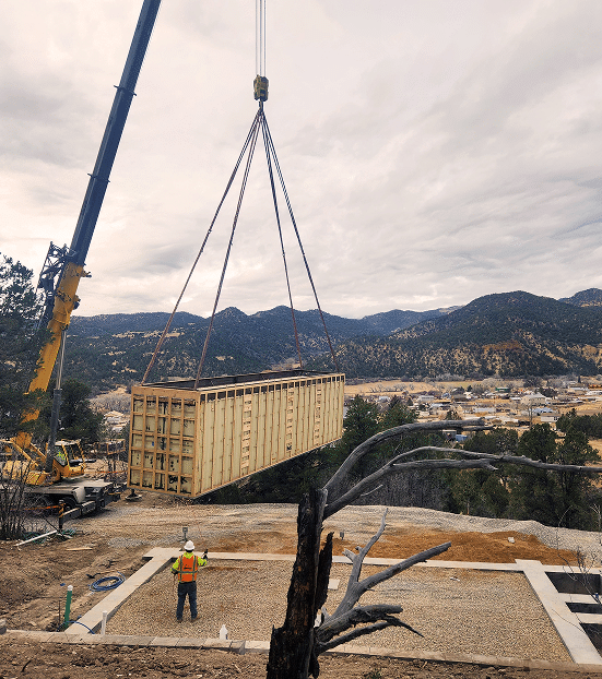 Construction crane lifting a wooden structure at a custom home building site, with a worker in safety gear overseeing the process against a mountainous backdrop.