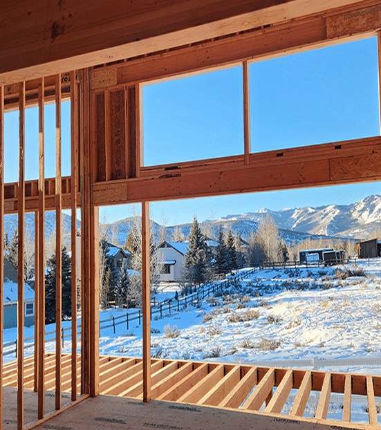 Framed interior view of a construction site showcasing windows with mountain landscape and snowy terrain, illustrating the construction phase of custom home building by Oak-n-Crete.